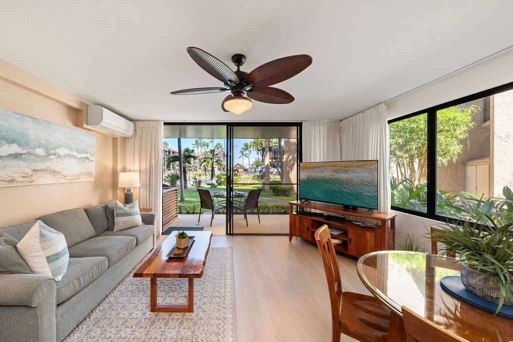 A modern living room with a sofa, TV, dining table, ceiling fan, and large windows.