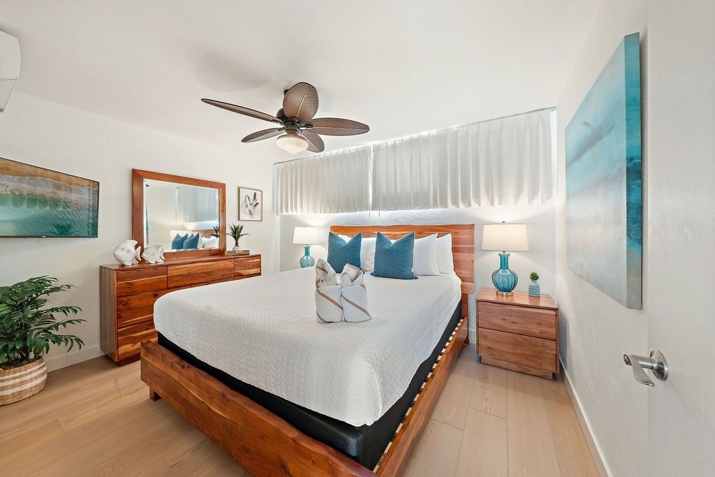 Een slaapkamer met een houten bed, een plafondventilator, nachtkastjes met lampen, een groot raam met gordijnen en een potplant.