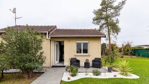 Une maison de plain-pied avec un toit en tuiles, un petit porche à l’avant doté de deux chaises, et un jardin où poussent différentes espèces de plantes.