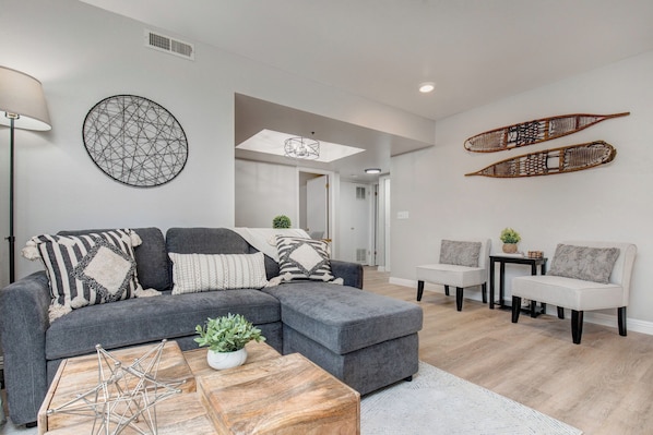 A modern living room with a grey sectional sofa, a coffee table, and two armchairs. There are decorative wall hangings and a small plant on the coffee table.