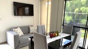 Een moderne woonkamer met een bank, eettafel en stoelen. Een flatscreen televisie is aan de muur bevestigd.