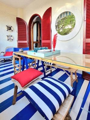 Una habitación con una mesa de vidrio, piso a rayas azul y blanco, contraventanas rojas y un espejo circular que refleja un árbol verde.