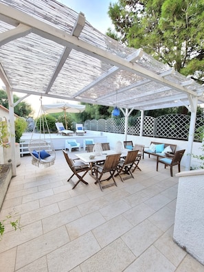 Un patio techado al aire libre con una mesa, sillas, una hamaca colgante y una cerca blanca.
