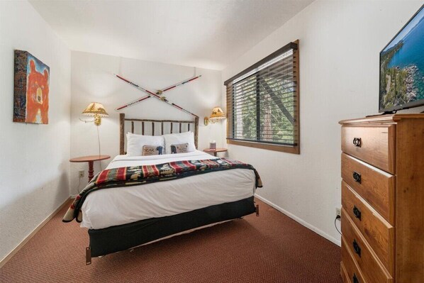 A bedroom with a bed, a wooden dresser, a TV, a window with blinds, and a painting on the wall.