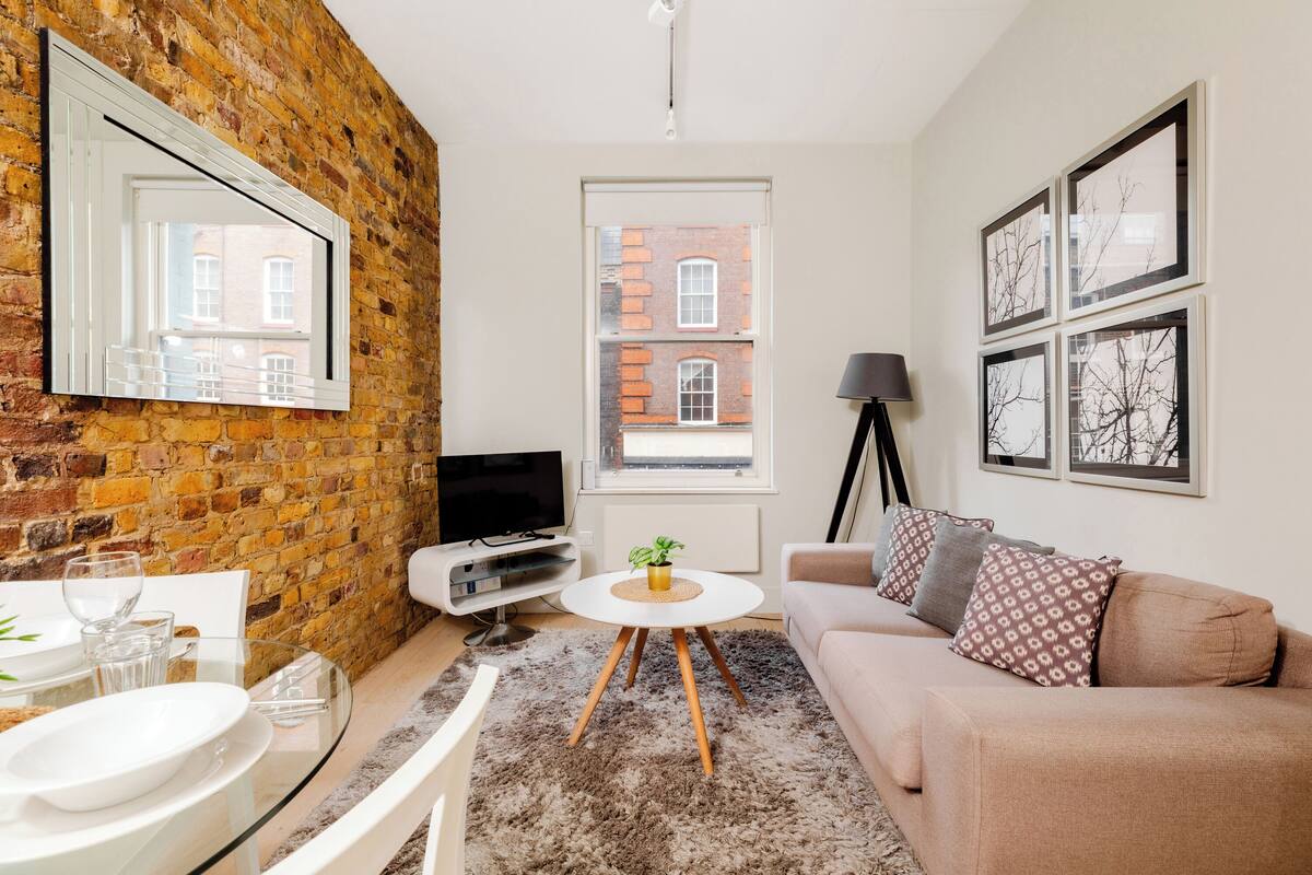 A modern living room with a brick wall, a sofa, a coffee table, a TV, and a window with a view of buildings.