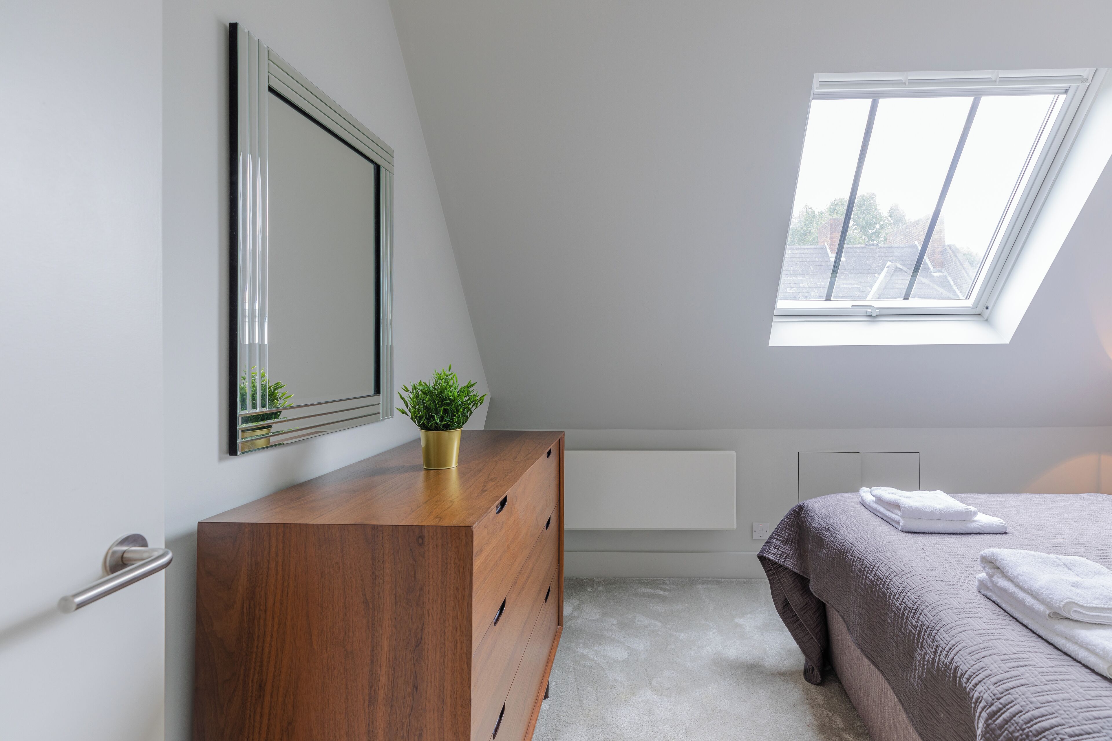 Een slaapkamer met een houten dressoir, een grote spiegel, een potplant, een bed met grijze dekbedovertrek en een dakraam.