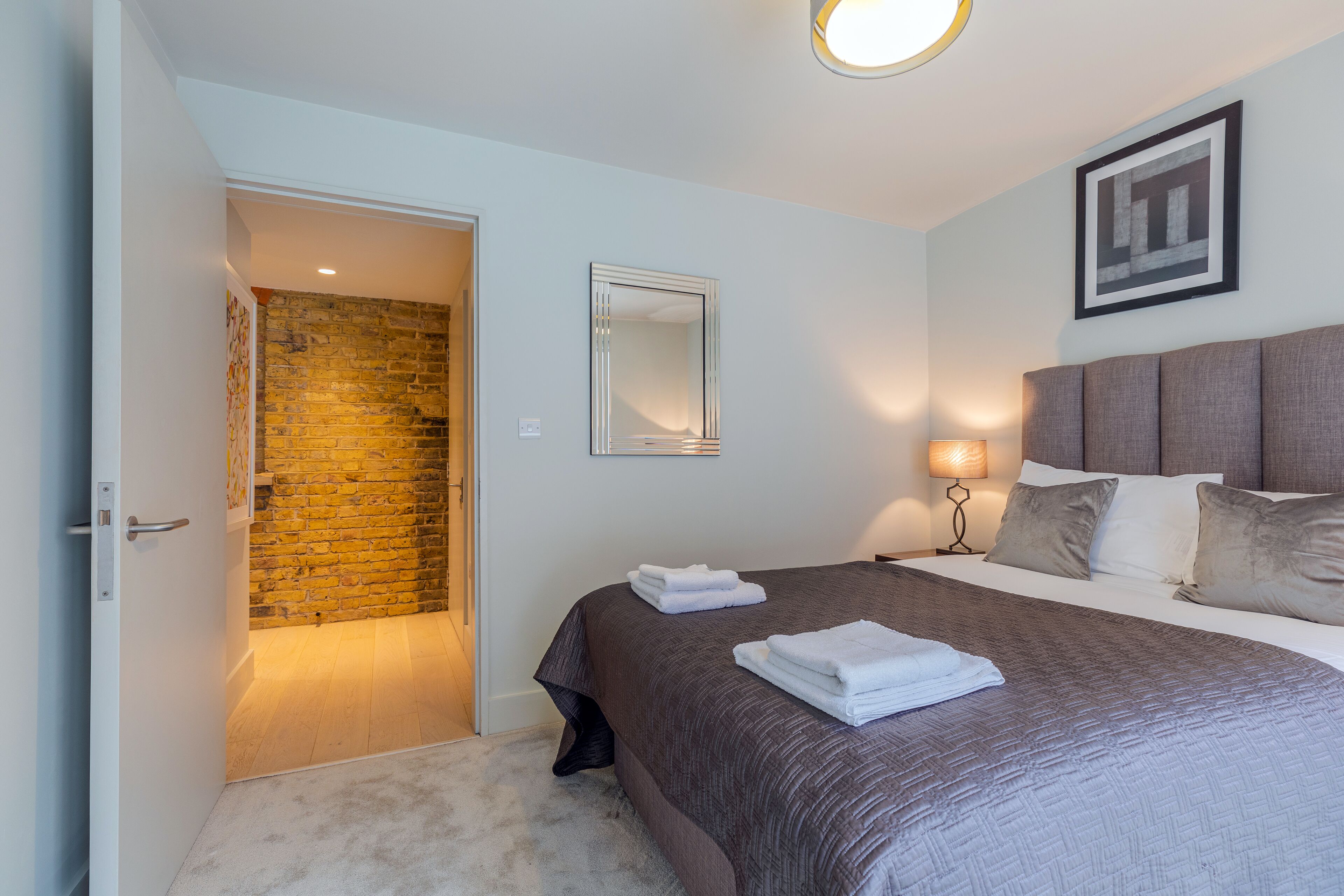 A hotel room with a bed, a nightstand, a lamp, a mirror, and a door leading to a bathroom.