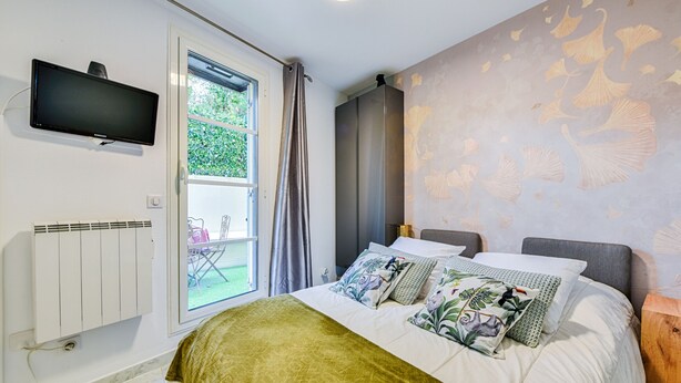 A bedroom with a bed, a TV mounted on the wall, a window with curtains, and a radiator. - Happyfew La Providence (Nice)