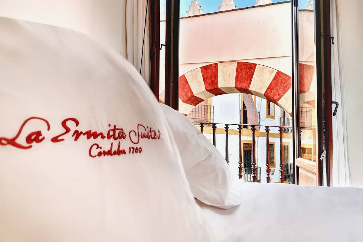 Habitación de hotel con cama, ventana con vistas a un edificio con un arco rojo y blanco a rayas, y almohada blanca con texto.
