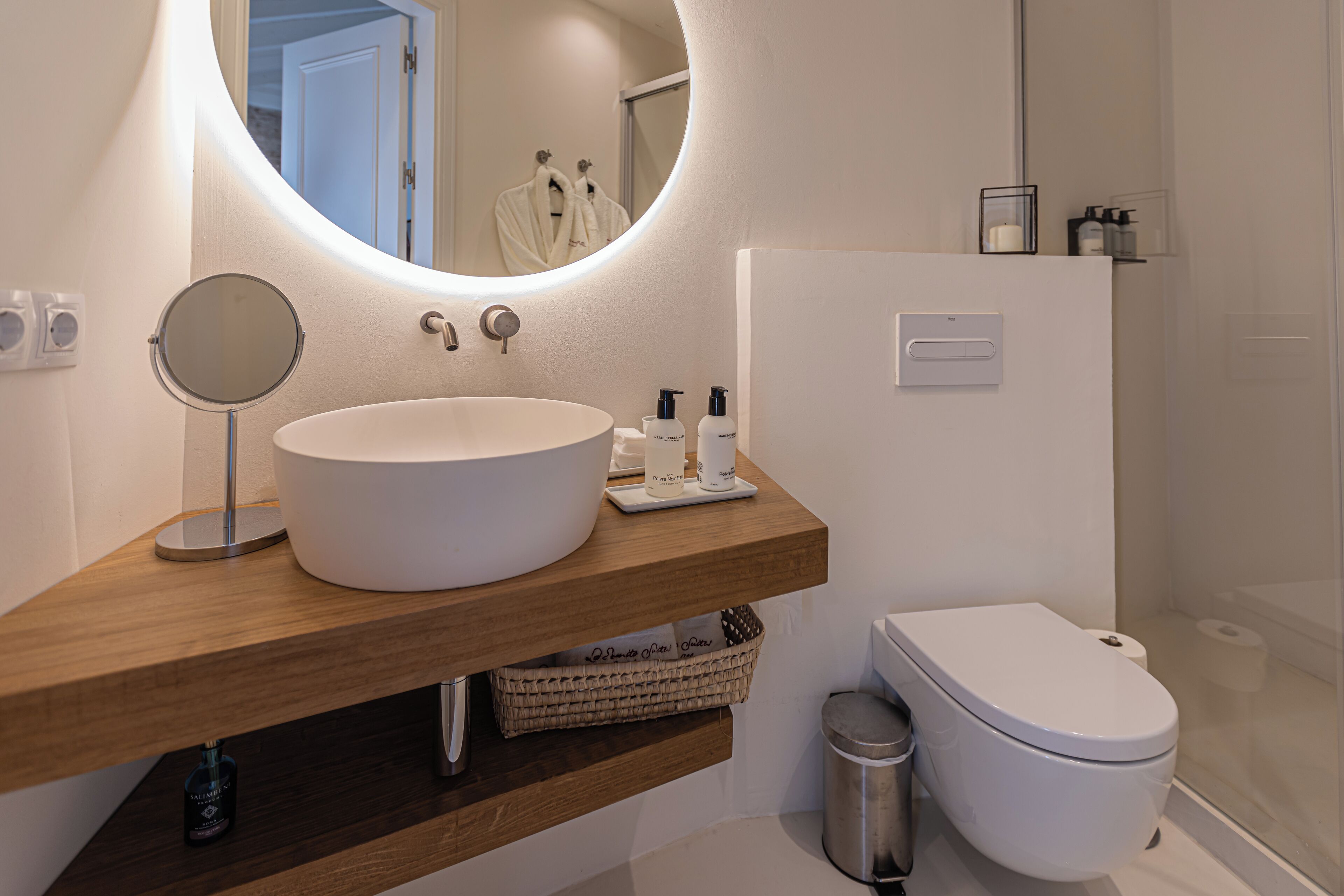 Un baño con un espejo redondo, un mueble de tocador de madera, un lavabo blanco y un inodoro blanco.
