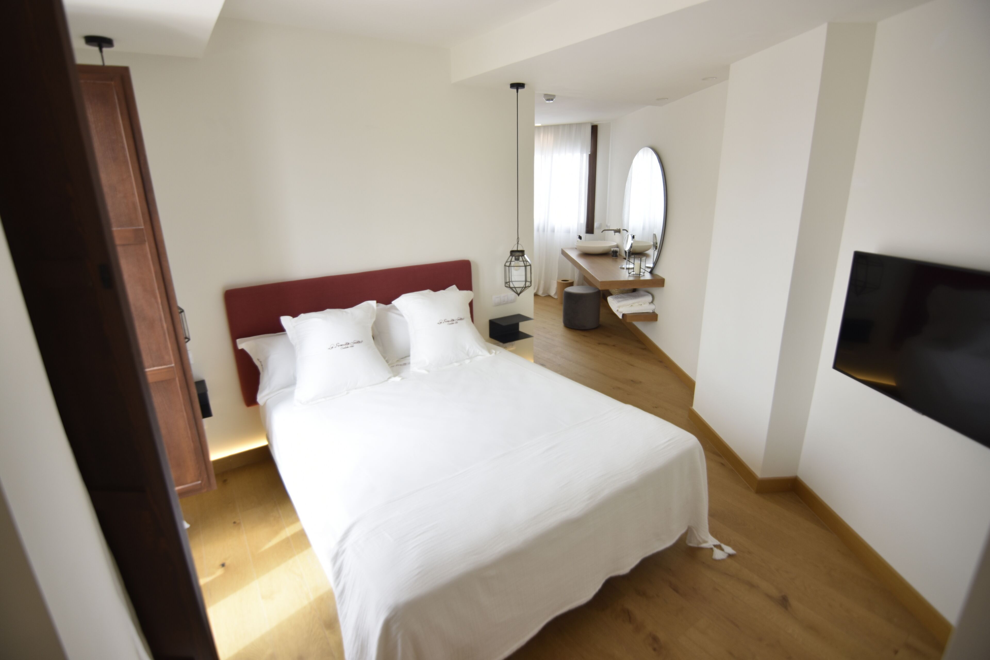 Un dormitorio con cama, suelo de madera, televisor y baño con lavamanos y espejo.