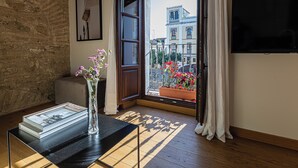 Ein Zimmer mit Holzboden, einem Glastisch mit Buch und Vase, einem Fenster mit Blick auf Gebäude und einem Balkon mit Blumen.