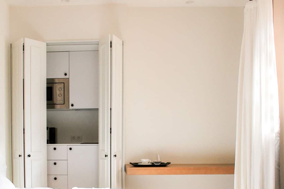 Un dormitorio con cama, armario abierto con electrodomésticos de cocina, estante de madera y ventana con cortinas.