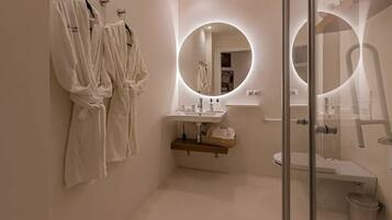 A modern bathroom with two white robes hanging on hooks, a round mirror with a light, a sink with a wooden shelf below, and a glass shower enclosure.