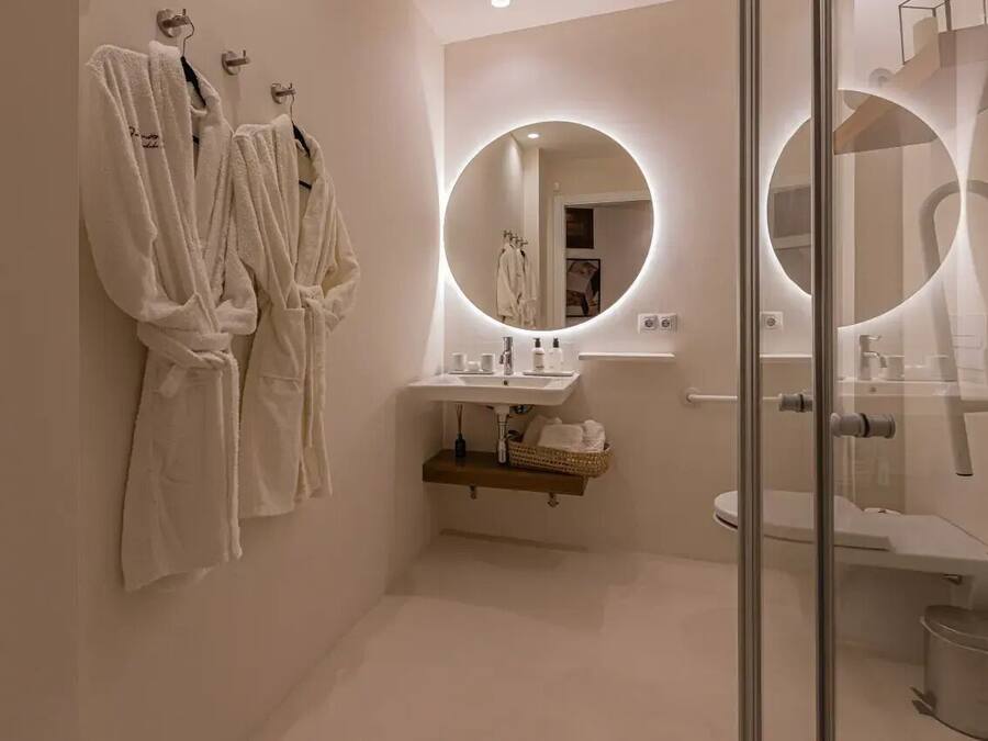 A modern bathroom with two white robes hanging on hooks, a round mirror with a light, a sink with a wooden shelf below, and a glass shower enclosure.