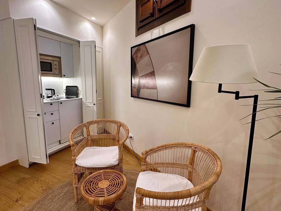 Una habitación con sillas de mimbre, una lámpara de pie, una zona de cocina y una obra de arte enmarcada en la pared.
