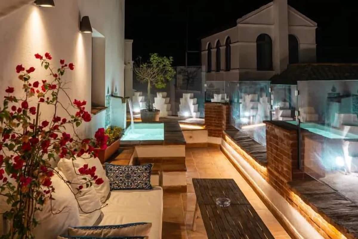 Un área de estar al aire libre con un sofá y una mesa, una planta en maceta y un edificio con fachada blanca y ventanas de arco.