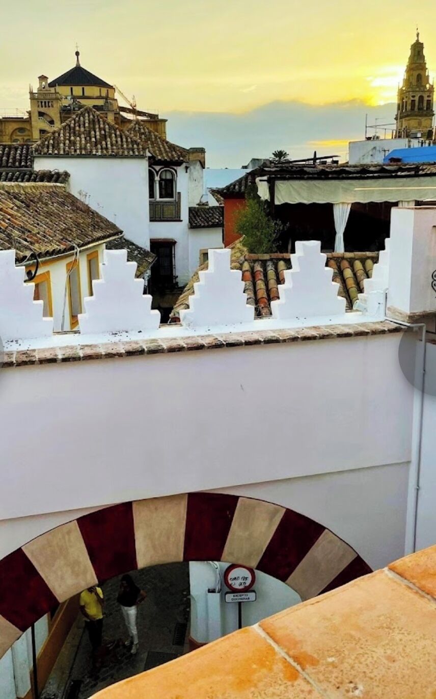 Vista de un edificio con fachada blanca y tejado de tejas, con vista a una calle con gente y un letrero que indica zona de no estacionamiento.