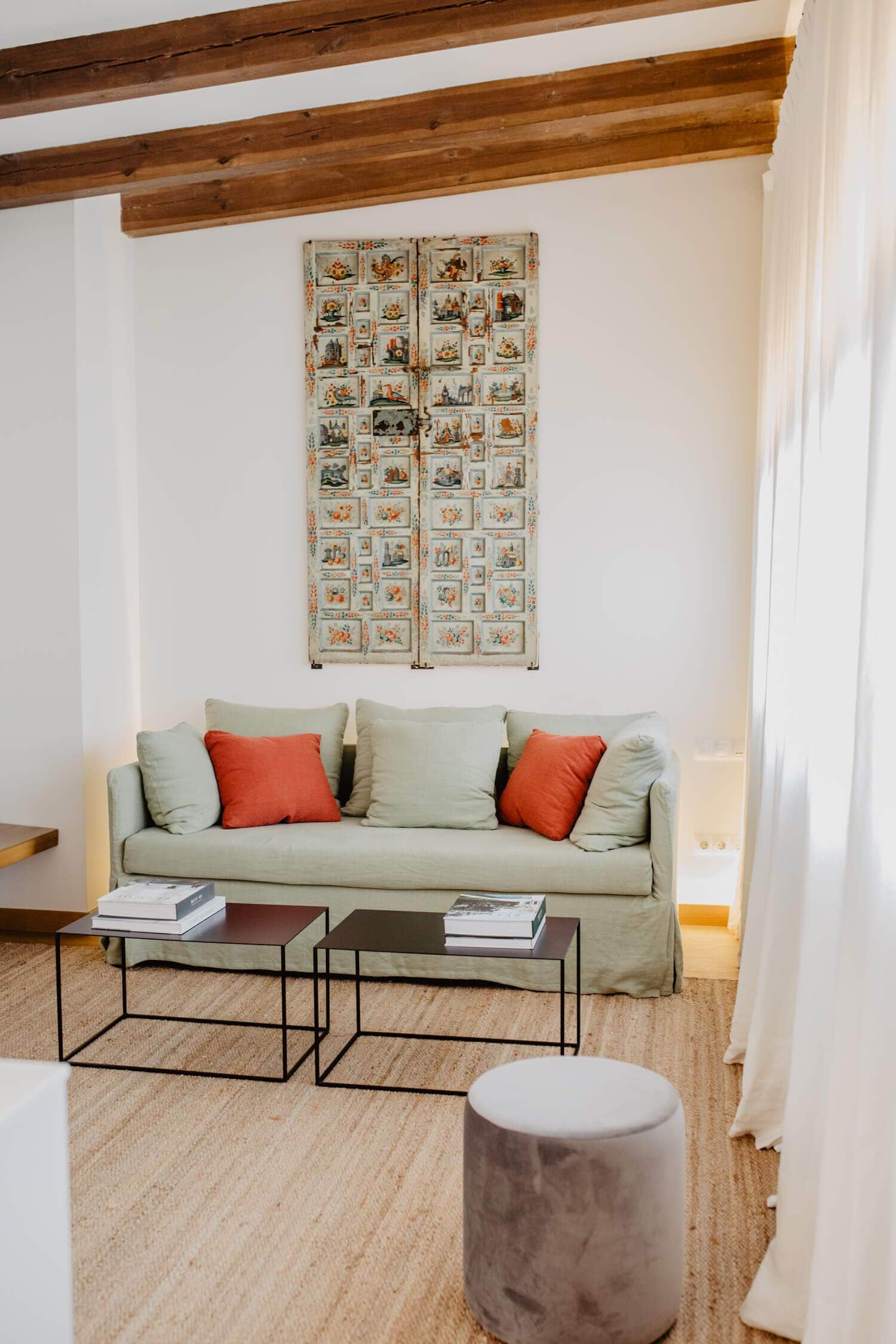 Un salón con sofá, dos mesitas auxiliares, un otoman redondo y una obra de arte en la pared.