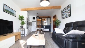 A modern living room with a black leather sofa, a wooden coffee table, and a flat-screen TV. There is a kitchen area with a refrigerator and a dining table.