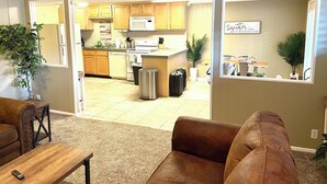 An open-plan living and kitchen area with a sofa, a small table, a trash bin, a suitcase, and a potted plant.
