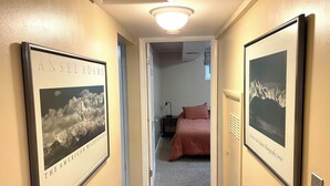 A hallway with framed photographs on the walls, a bed in the adjacent room, and a ceiling light.