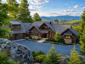 A large, multi-story house with a stone foundation, multiple chimneys, and a spacious front yard with a gravel driveway. - Twin Boulder Lodge at Eagles Nest: big views, outdoor fireplaces, hot tub, arcade (Banner Elk)