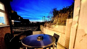En terrasse med et rundt bord og stole, en lille sort genstand på bordet, og udsigt til byens skyline i skumringen.