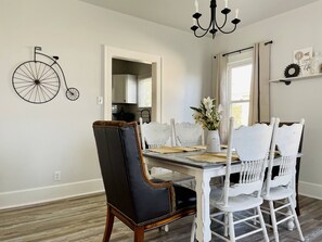 Un comedor con una mesa dispuesta para cuatro personas, rodeada de sillas blancas con patas de madera. Una silla negra y marrón está ubicada al frente de la mesa. Se observa una bicicleta decorativa negra fijada en la pared izquierda.