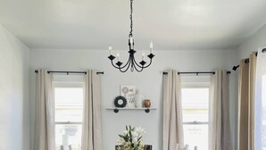 A dining room with a chandelier, white chairs, a dark upholstered chair, a wooden floor, and a window with curtains. - The Chic Shack | Centrally Located 2BR/1BA Home (Evansville)