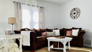 A living room with a brown sofa, a white side table, a round coffee table, and a wall clock. - The Chic Shack | Centrally Located 2BR/1BA Home (Evansville)