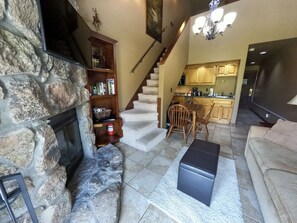 A living room with a stone wall, a fireplace, a bookshelf, a staircase, and a kitchen in the background.