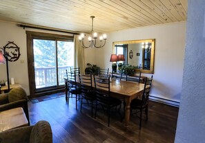 A dining area with a wooden table and chairs, a chandelier, and a large window with a view of trees.