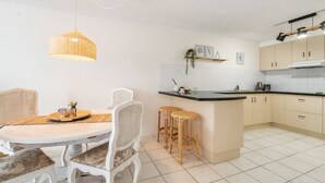 Une cuisine moderne avec des meubles blancs, un îlot central et un coin repas agrémenté de chaises en osier et d’une table blanche.
