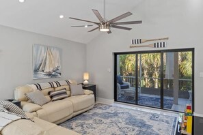 Un salon avec un canapé beige, un ventilateur de plafond, une grande fenêtre donnant sur des palmiers et un tapis à motifs.