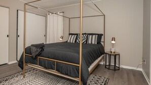 A four-poster bed with a black bedspread and pillows, a small bedside table with a lamp, and a patterned rug on a wooden floor.