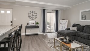 A modern living room with a grey sectional sofa, a dining area with a marble countertop and black chairs, and a large window with curtains.