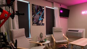 Ein Zimmer mit zwei Stühlen, einem kleinen Tisch, einem wandmontierten Fernseher und einer Neonreklame.