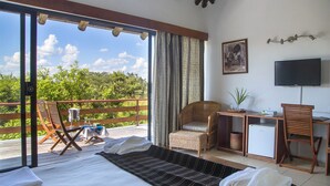 Una habitación con cama, escritorio, silla, mesita y televisor. Cuenta con un balcón con sillas y mesa, y vistas a árboles y cielo.