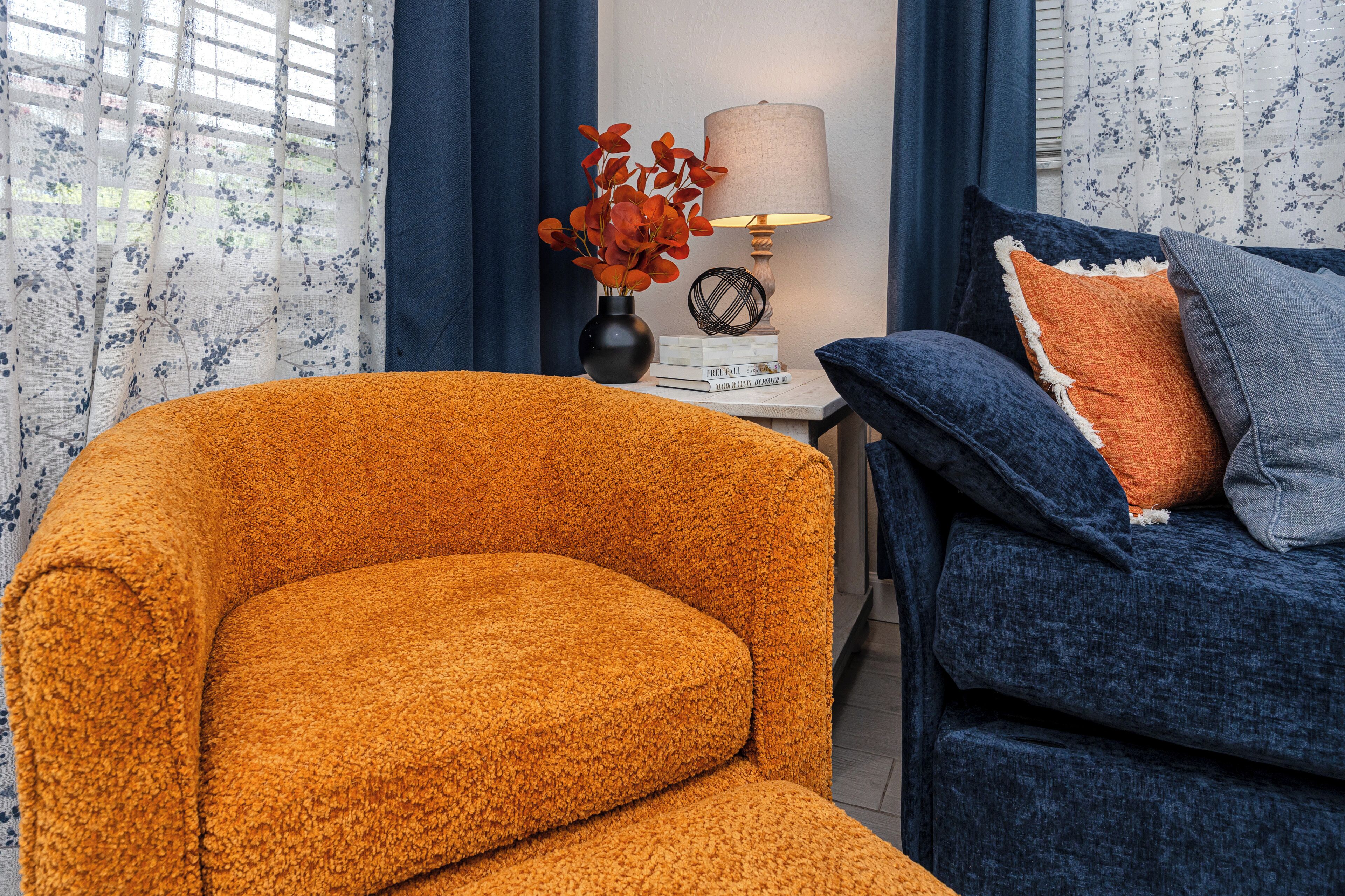 A room with a mustard-colored armchair, a dark blue sofa with an orange pillow, a lamp, and a vase with red flowers on a side table.