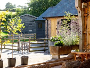 A rustic outdoor seating area with wicker furniture, a concrete planter with flowers, and a wooden fence. - Frith Green (Bromyard)