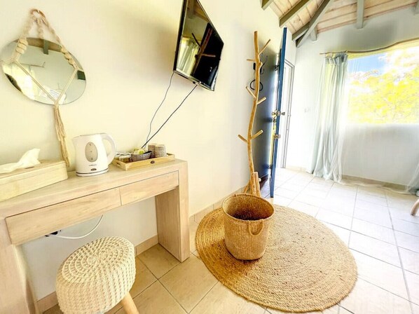 Ein Zimmer mit Schreibtisch aus Holz, einem runden Spiegel, einem an der Wand befestigten Fernseher, einem gewebten Korb und einem Fenster mit Vorhängen.