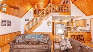 A spacious living room with a high wooden ceiling, a staircase leading to an upper level, and a kitchen area visible through an open doorway.