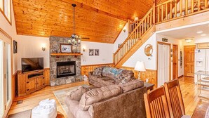A spacious living room with a stone fireplace, wooden ceiling, and a staircase leading to an upper level.