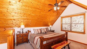 A bedroom with a wooden ceiling, a bed with a wooden headboard, a nightstand, a bench, and a window with a view of a snowy landscape.