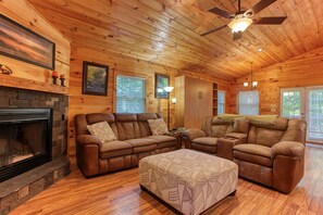 A cozy log cabin interior with a fireplace, leather sofas, and a ceiling fan. - Experience mountain living at The Fiddlers Green (Bryson City)
