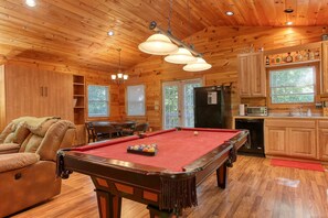 A wooden cabin interior with a pool table, a sofa, a kitchen area, and a dining table.