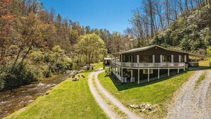A two-story cabin with a porch, surrounded by a forest and a stream.