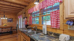 Interior de uma cabana de madeira com cozinha, cortinas xadrez, pia e área de refeições com um banco.