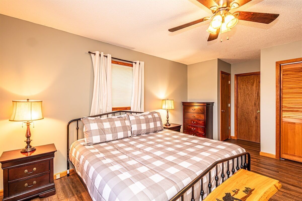A bedroom with a bed, two bedside tables, a nightstand, a dresser, a ceiling fan, and a window with curtains.